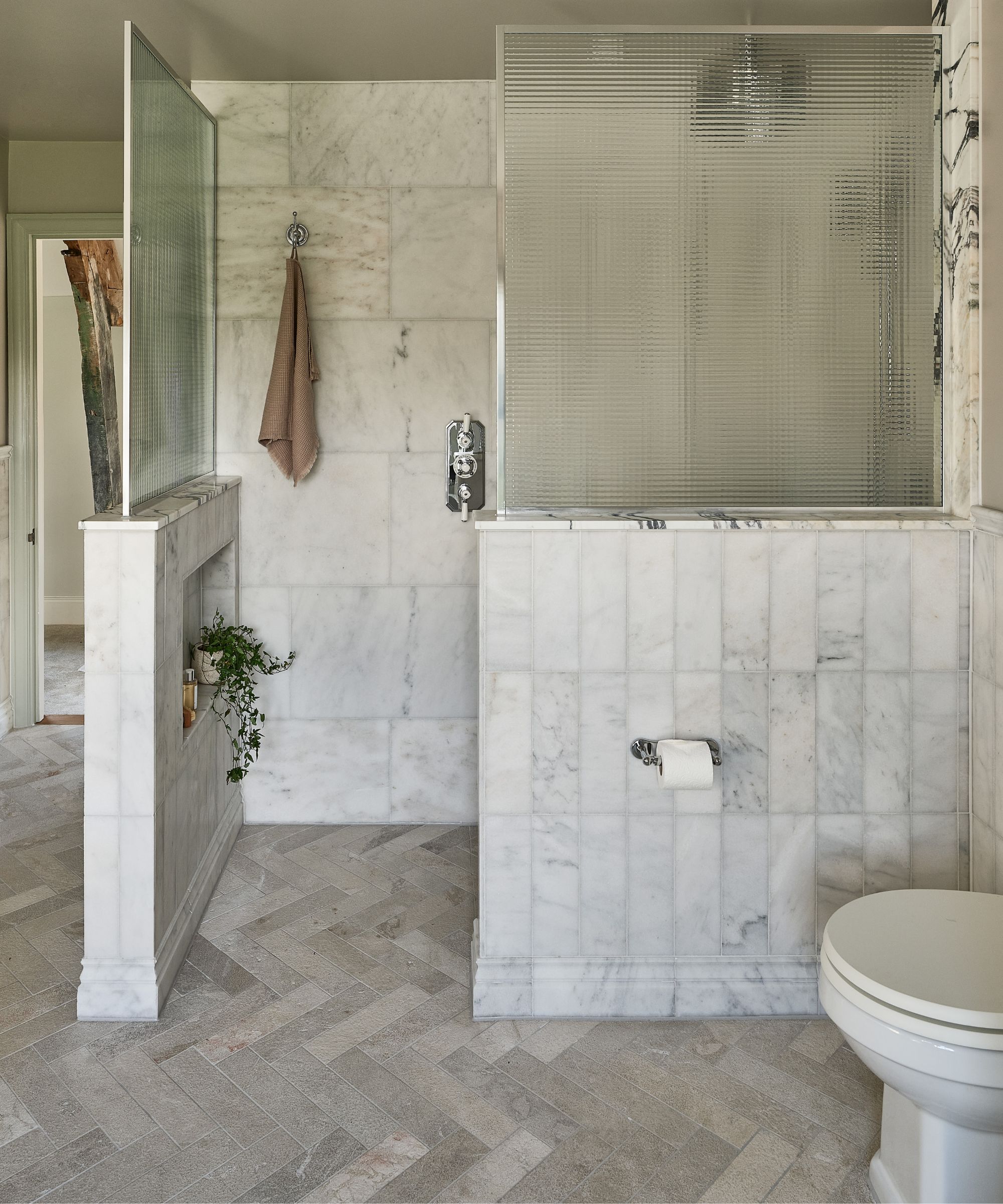 Marble-tiled bathroom with herringbone floor, walk-in shower behind ribbed glass, built-in shelves, and toilet set against soft neutral walls.