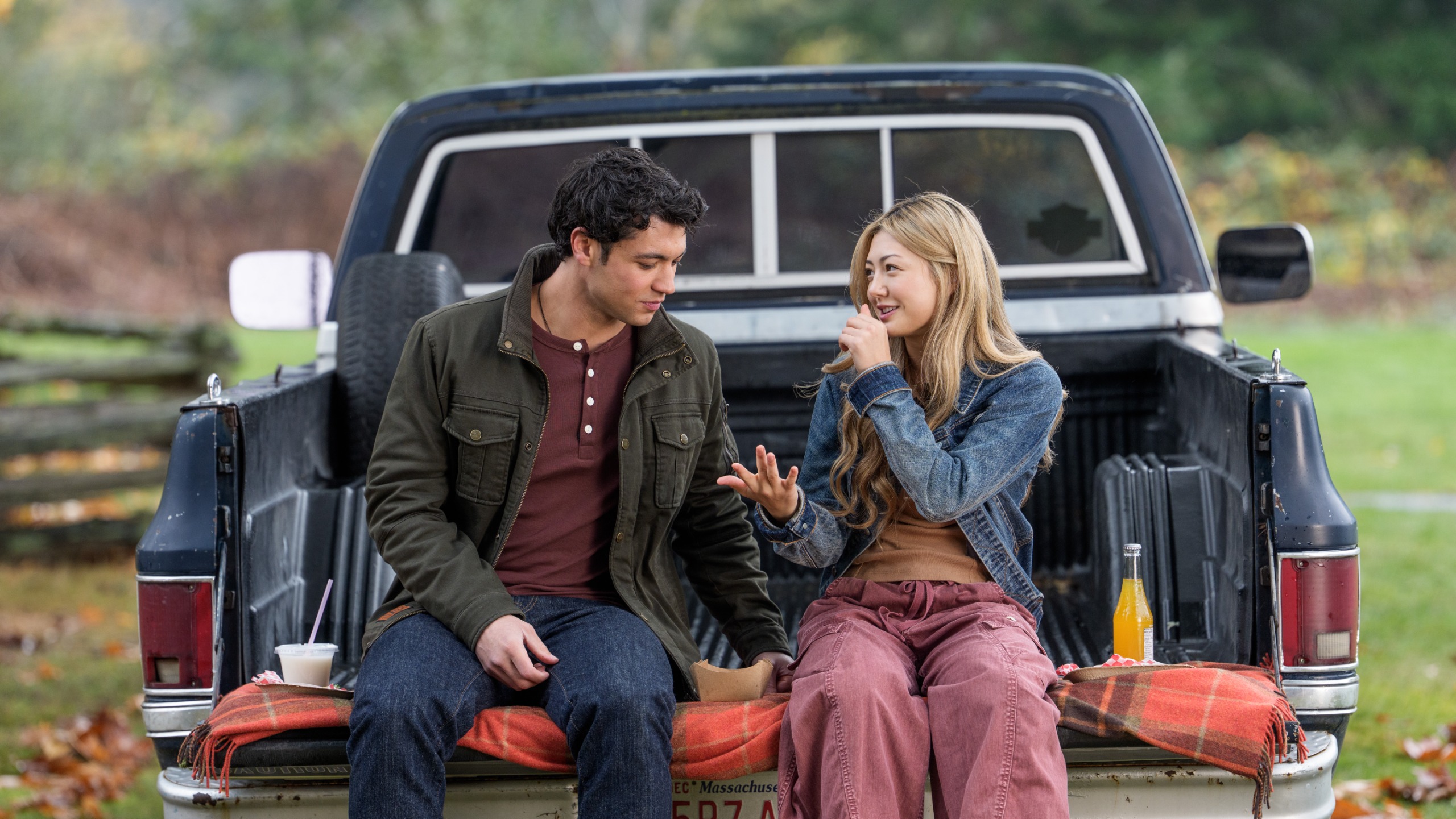 A teenage boy and girl (Kyle Clark as Cam Dillinger and Miku Martineau as Teagan Tao) have a picnic on the bed of a pickup truck, in Netflix's 'Crew Girl'.