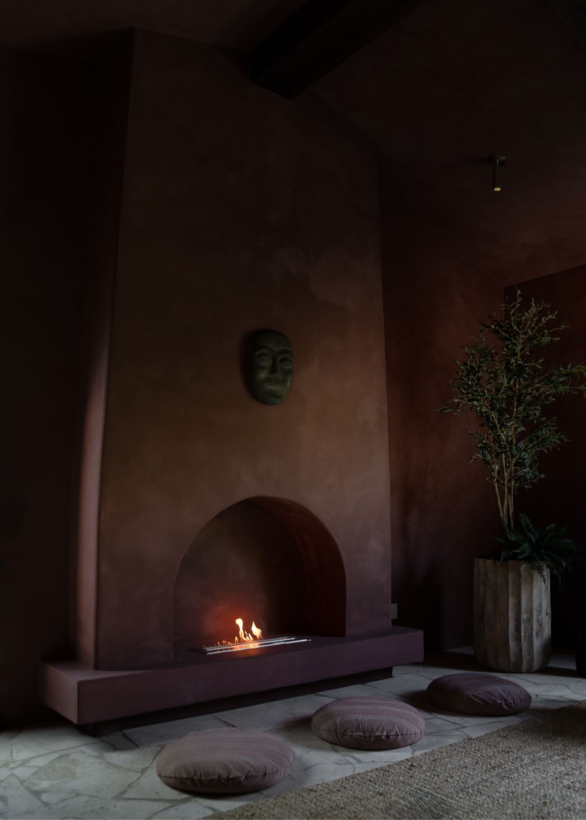 A dark brown meditation room with a potted tree, an arched fireplace, and round purple floor cushions