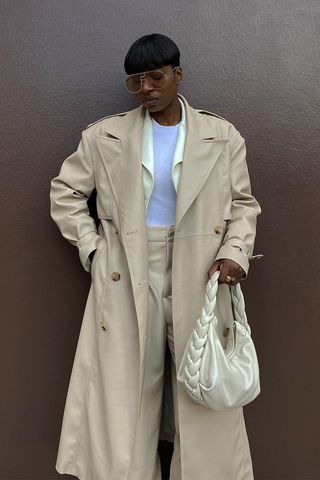 spring trench coats shown in a photo of a French woman standing outside against a brown wall wearing brown aviator sunglasses, a tan trench coat over a creme leather blazer, white t-shirt, tan trousers, and a creme handbag