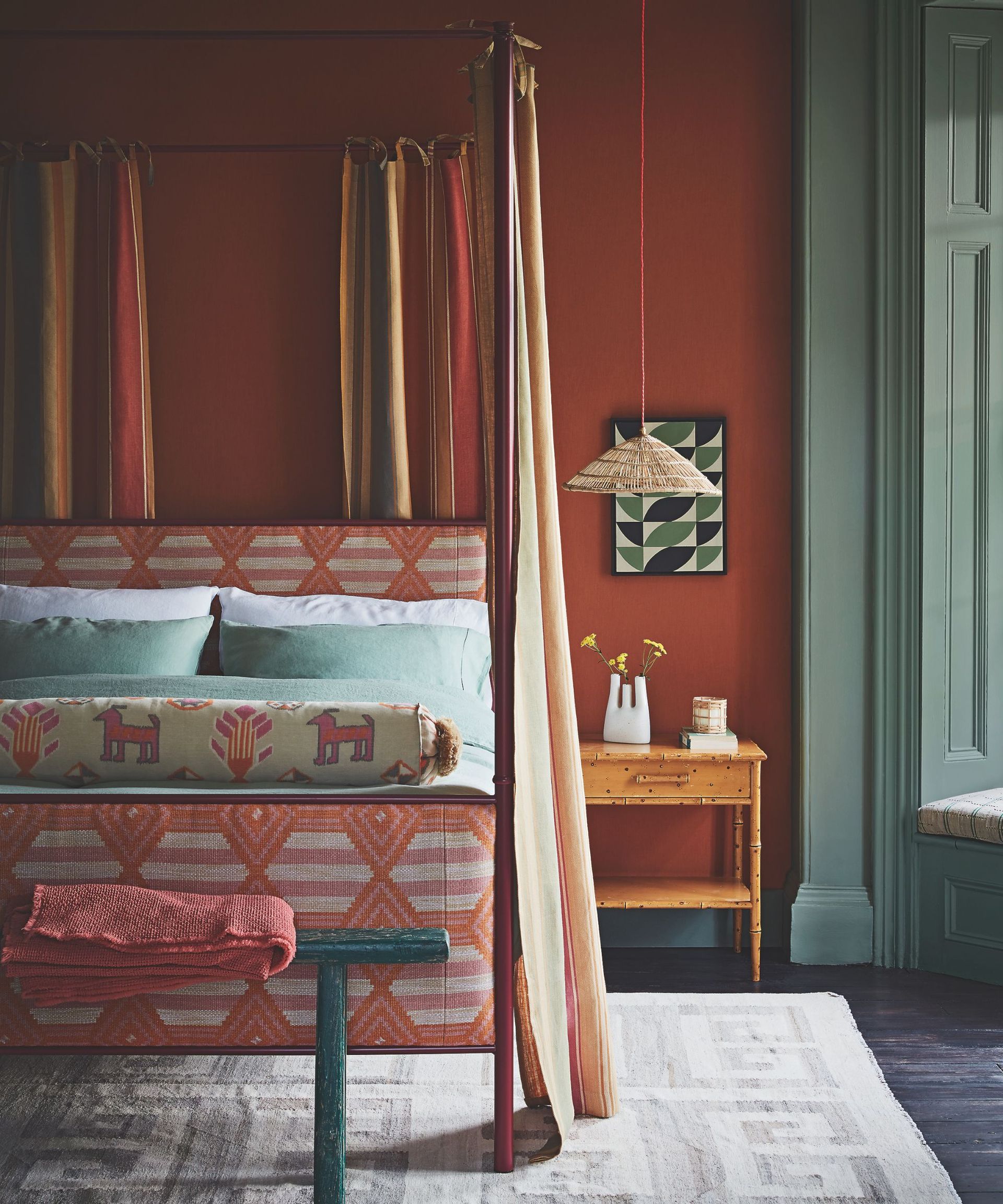 A burnt orange bedroom with sage green windows, a orange four poster bed with cream, and orange curtains, a green wooden bench at the end of the bed, and a cream patterned rug beneath. On the wall behind is a green and black framed artwork and a wooden nightstand with a white vase.