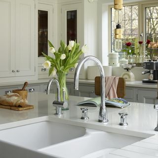 a white kitchen sing with 2 mixer taps on a kitchen isalnd with a vase of flowers and a chopping board with fresh bread