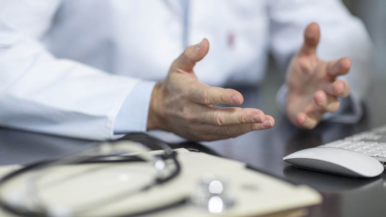 Doctor gestures with his hands above desk that holds files and a stethoscope.