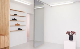 The Packyard store is all white, with a glass separator diving the room. To the left, we see light wood shelves with shoes on them. To the right, we see a black vase with flowers in it.