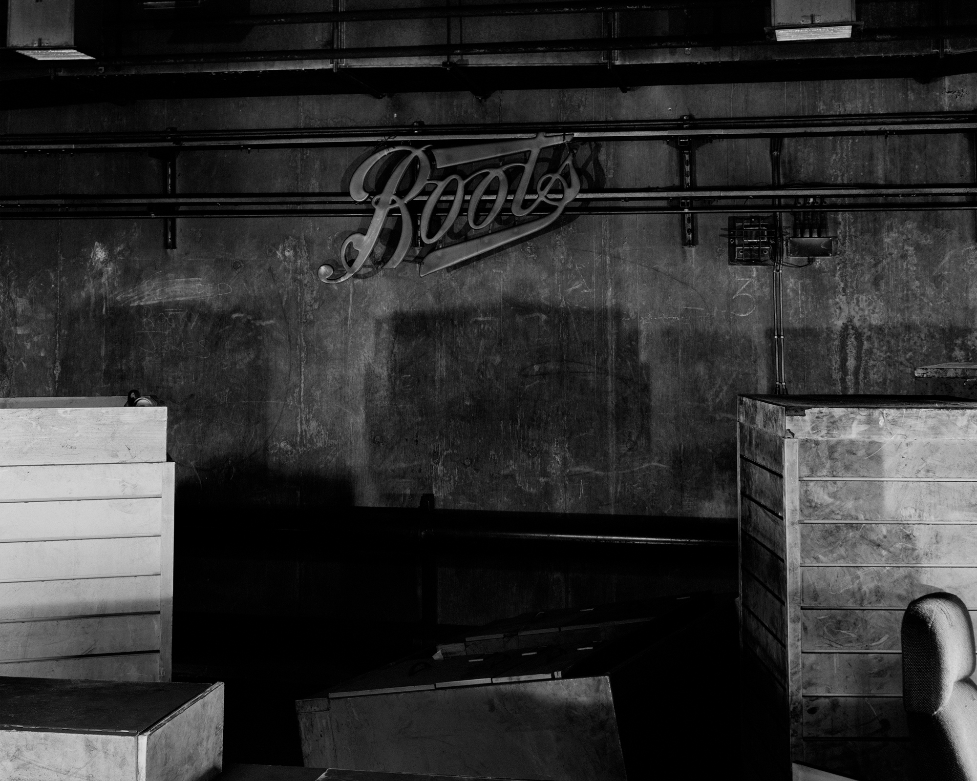 Black and white photograph of a dim, gutted former shop interior with an old neon "Boots" sign still fixed to the back wall.