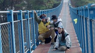 three people (lee kwang-soo, yoo jae-suk, and kwon yuri) sit and kneel on a bridge, in 'the zone survival mission'