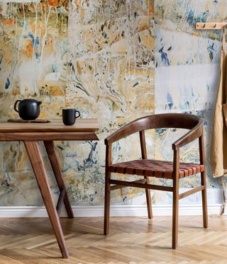 View of a space with multicoloured marble patterned wallpaper, wood flooring, a wooden chair and a wooden table with a small black teapot and mug on top