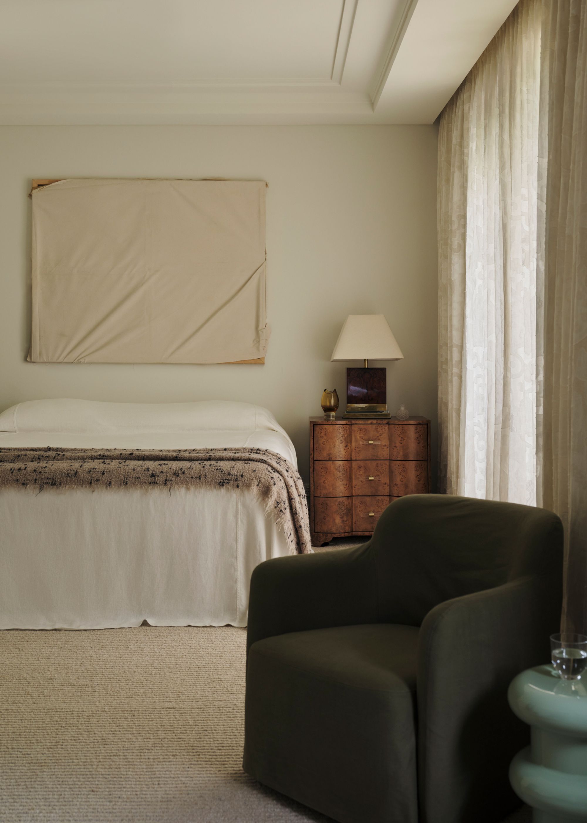 A minimalist bedroom with a burl nightstand, a table lamp, a minimalist bed, a framed cloth, a green armchair, and a stacked side table