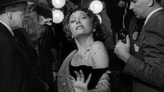 Gloria Swanson, in makeup and dress, walking in front of police cameras in Sunset Boulevard