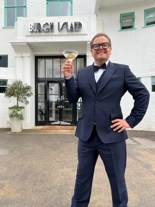 Alan Carr stands outside the main entrance to Art Deco-style Burgh Island Hotel. He is wearing a 1920s-style suit complete with bow tie, and holding up a fluted wine glass by the stem.