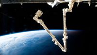 a multi-segmented arm extends from a cylindrical space station module. the blue-and-white curvature of earth is visible below