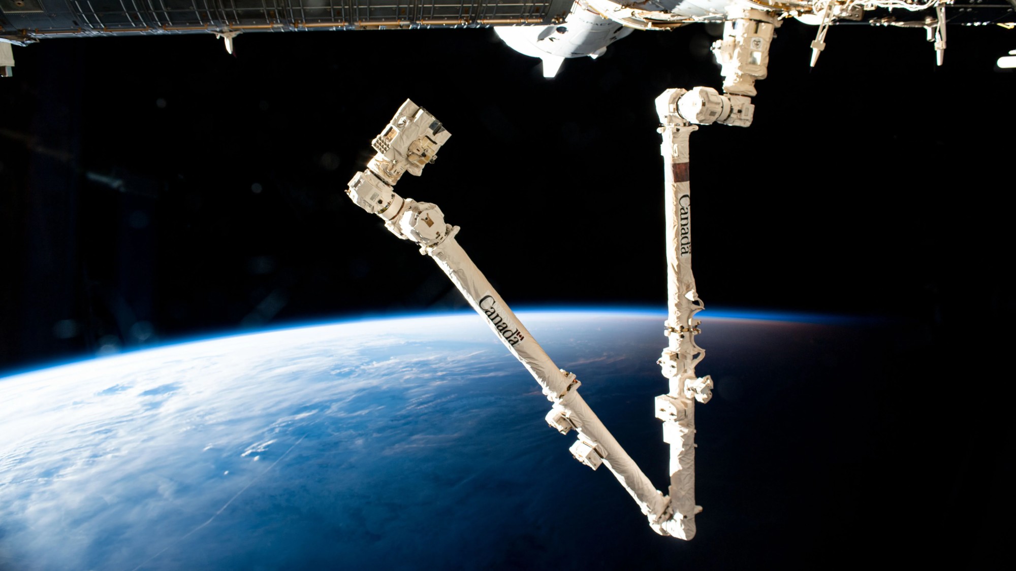 a multi-segmented arm extends from a cylindrical space station module. the blue-and-white curvature of earth is visible below