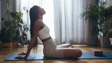 A woman relaxes in a seated position on a yoga mat at home