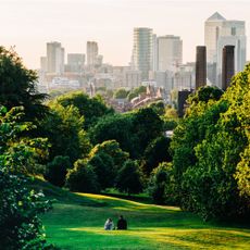 Two people sit in a tree lined park in front of a city skyline