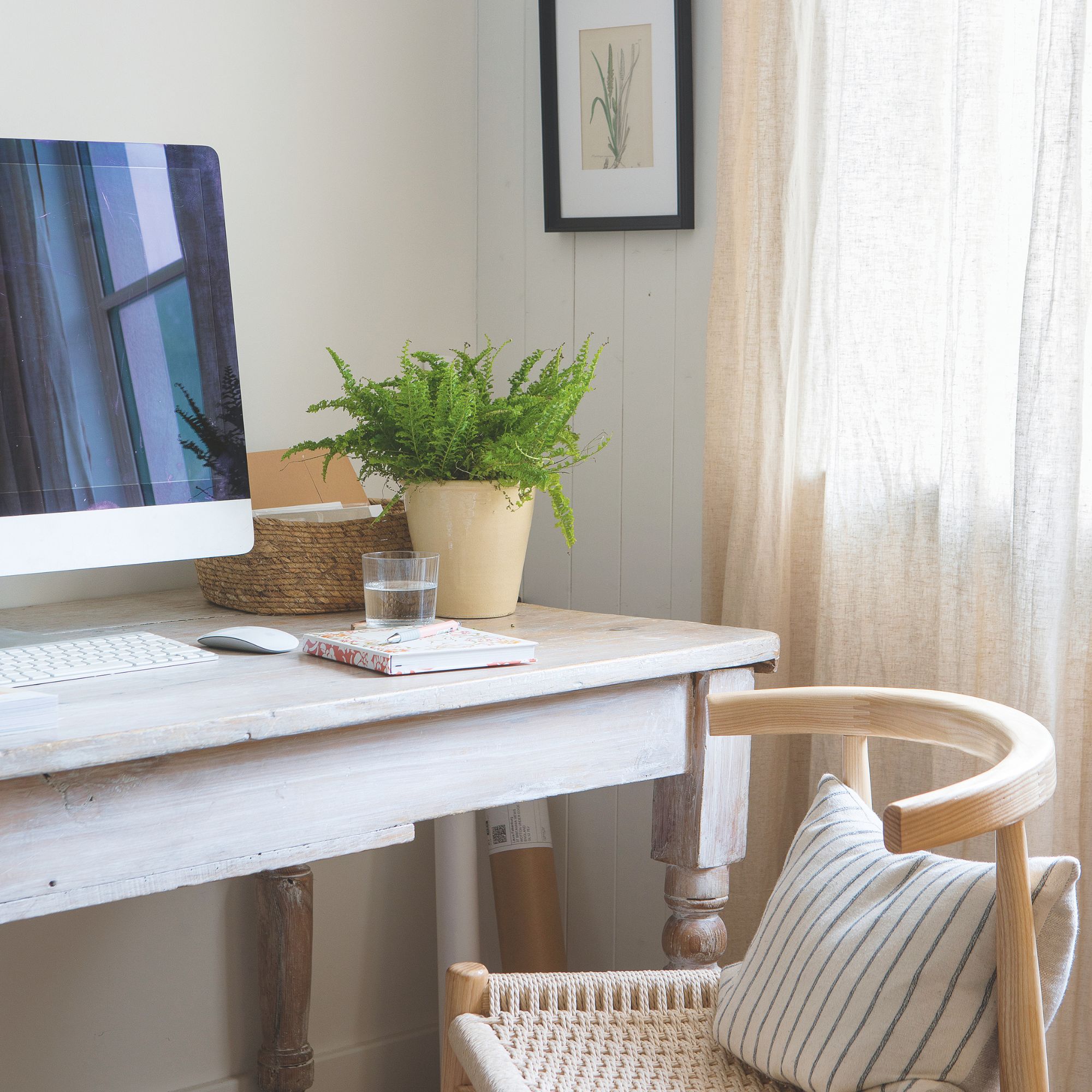Wooden desk with a desktop computer on it, with a wooden chair sitting in front of it, and linen curtains to the side