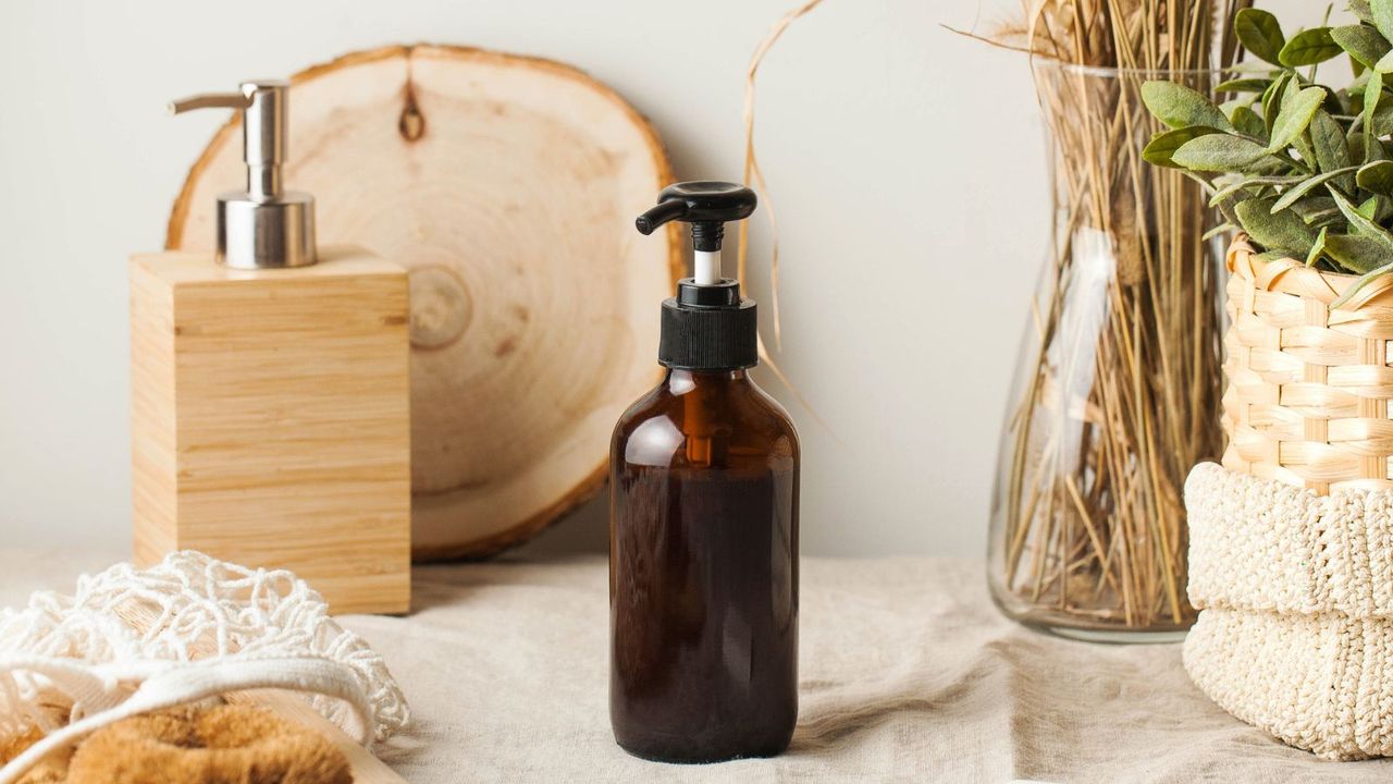 A dark brown glass soap dispenser on top of a beige cloth, with a wooden board, dried gras, and a green plant behind it