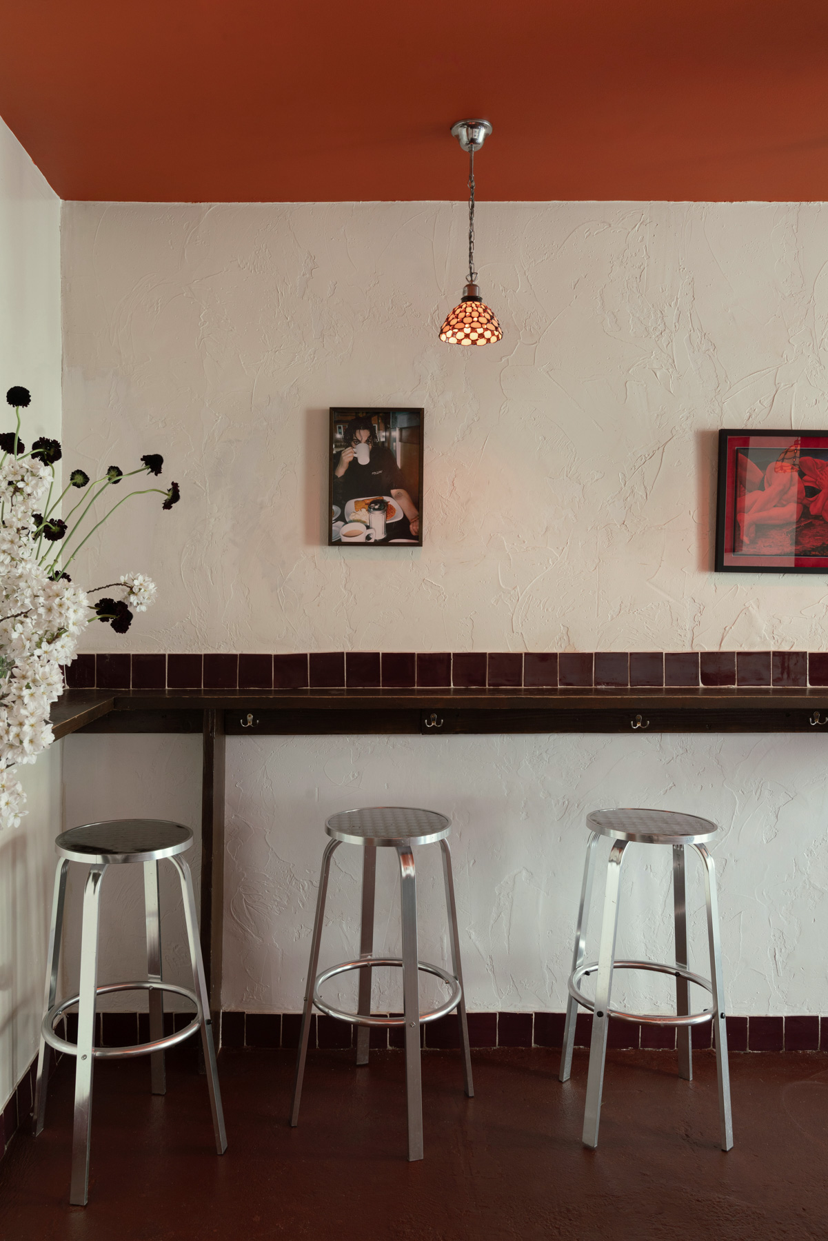 The interiors of a bar, with terracotta red ceilings, chrome and wooden furniture, framed artworks, and pendant lights in orange glass.