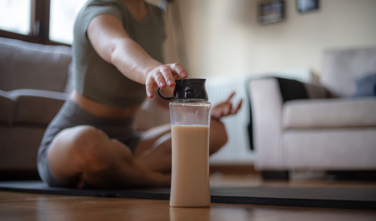 Woman&#039;s hand reaches for a chocolate protein shake in a see-through bottle. She is sitting cross-legged on the floor with couches behind her