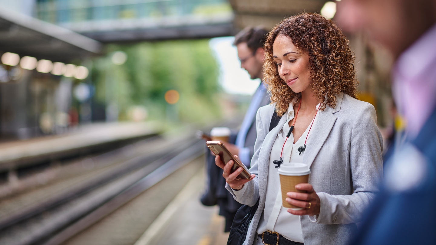 Woman standing on train platform holding her phone and a coffee