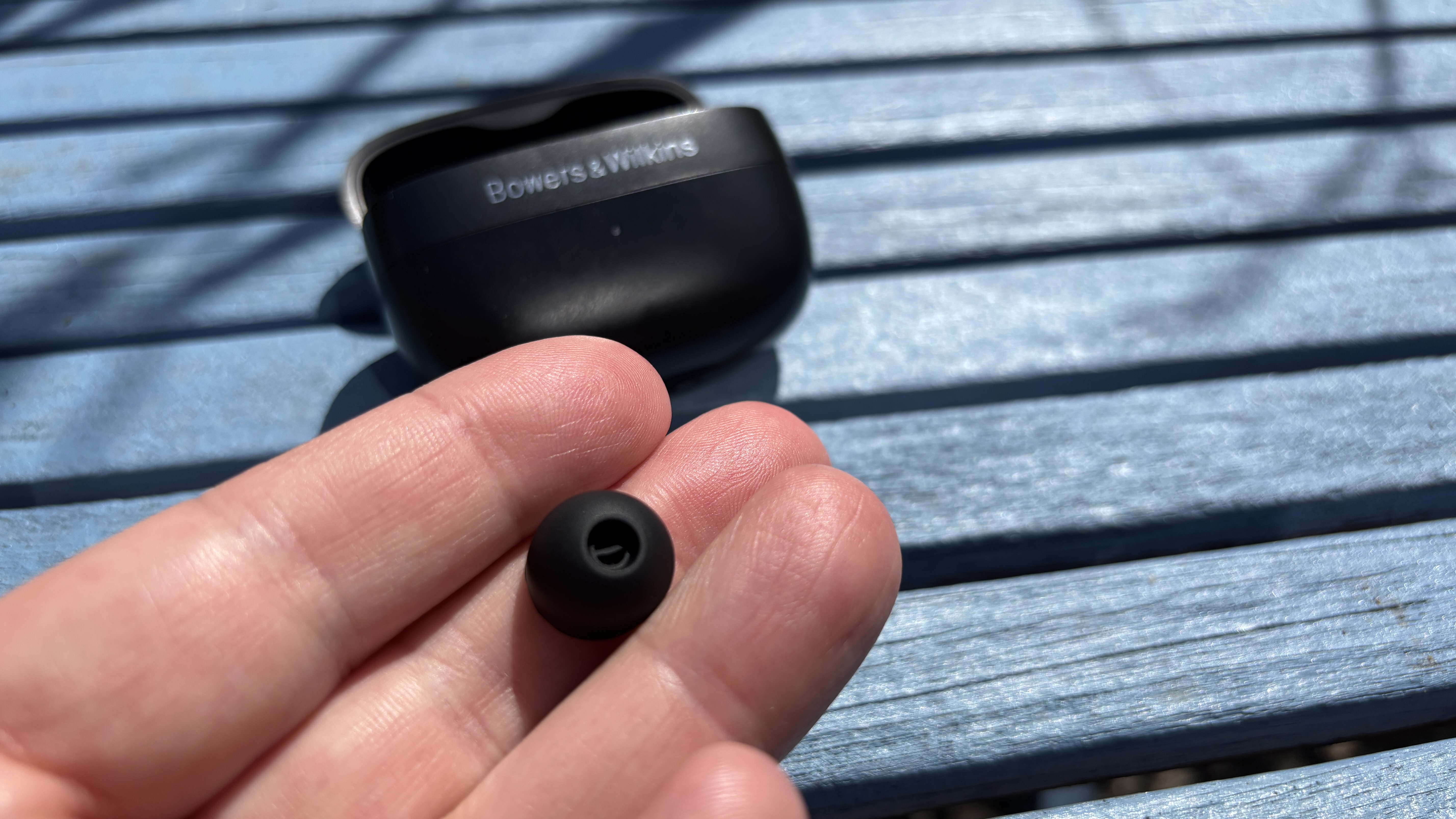 Bowers & Wilkins Pi8 wireless earbuds out of their case, placed on a blue table, with a detached ear tip being held in a man's hand