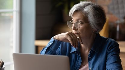 An older woman looks out the window, appearing to be thinking, while sitting in front of her laptop.