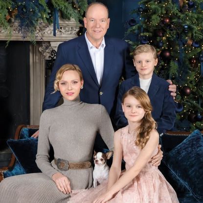 Princess Charlene wears a long taupe dress with a pleated skirt, long sleeves, and a turtle neck, and poses next to a burning fireplace and a Christmas tree with her pet dog, her twin children, Princess Gabriella and Prince Jacques, and husband Prince Albert