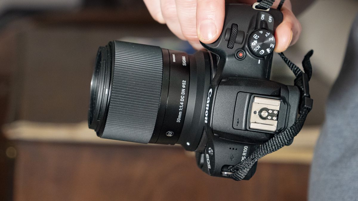 Sigma 30mm F1.4 DC DN Contemporary lens in the hand attached to a Canon EOS R100
