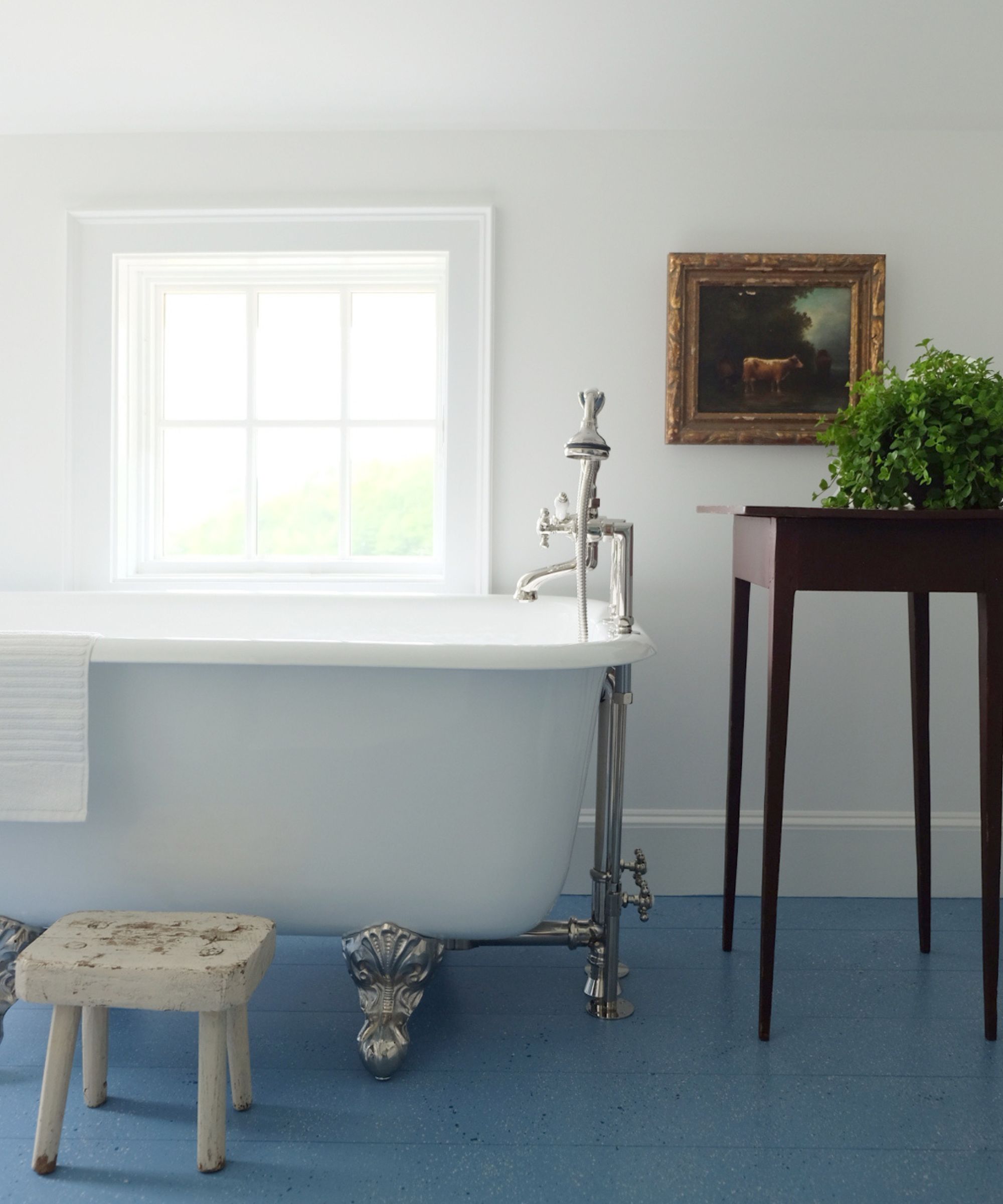 a white bright bathroom with a small window, freestanding bathtub, antique stools and a blue painted wooden floor