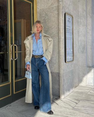 Elsa wears wide-leg jeans, black loafers, a Gucci belt, blue button-down shirt, beige trench coat, and white hermes bag.