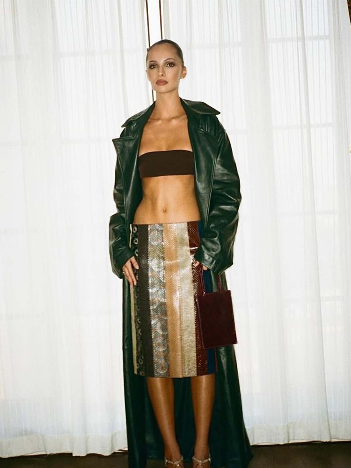 Pia Mance wearing a green trench coat, bra top, and printed striped skirt.