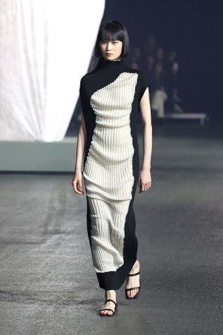 A female model wearing a long striped black and brown dress and black high heel shoes walking down a runway.