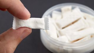 A close-up image of a nicotine pouch being held between someone's finger and thumb. Behind is a tub containing lots of nicotine pouches.