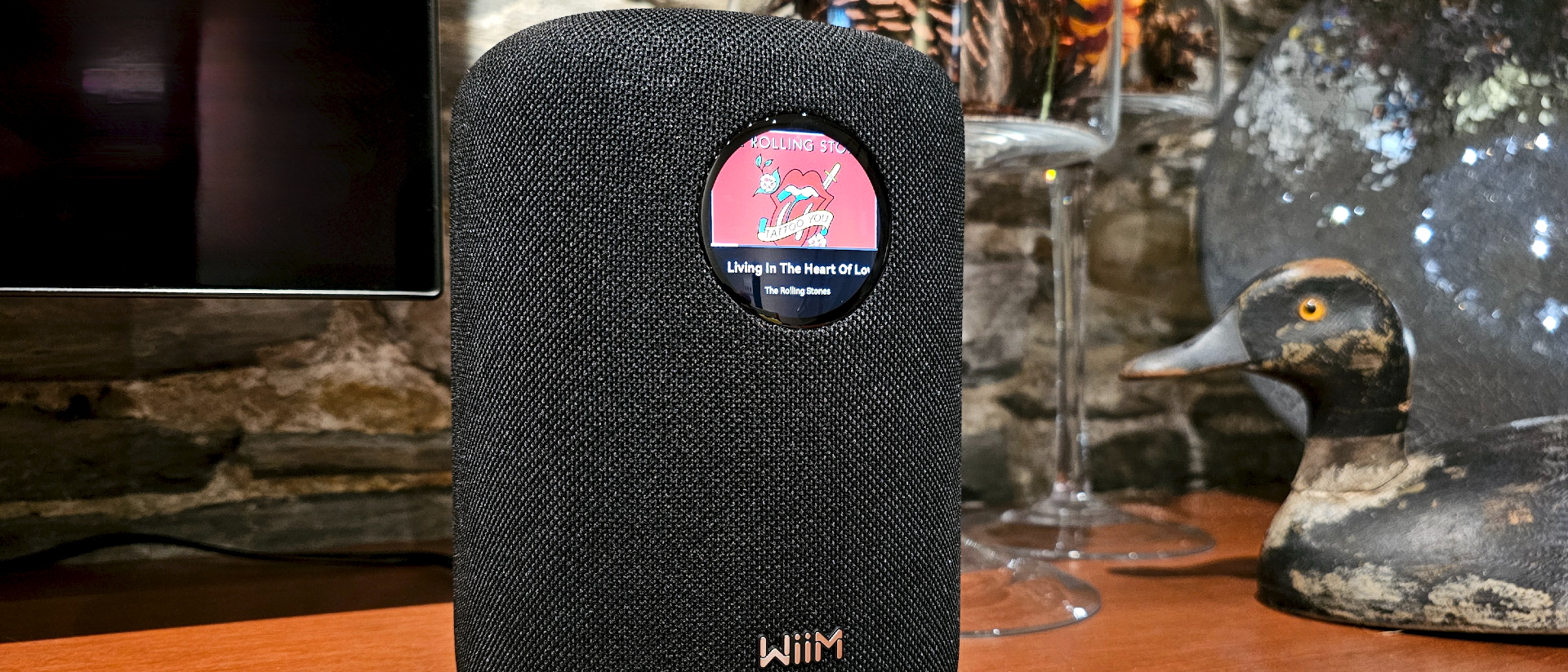 WiiM Sound on a wooden varnished table, with wine glasses and a TV in the background