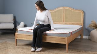 A woman sits on the end of a thin mattress in a bed frame in a bedroom. As the mattress has a small build, it lacks some support and cushioning