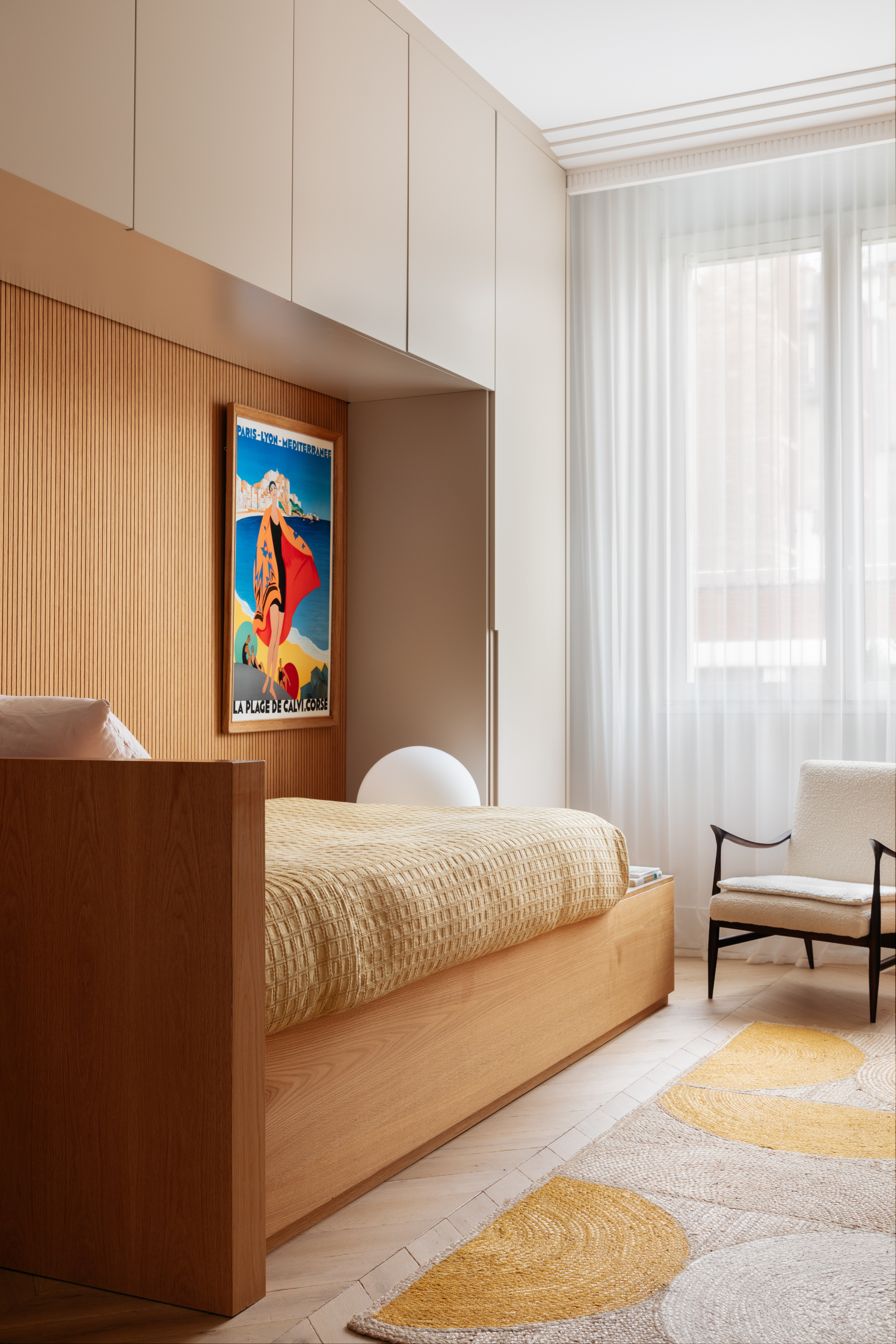 a chic kids room idea for an older child with a built in bed within built-in wardrobe all around it with a neutral color scheme and sheer white curtains with a white chair in front of the window and a piece of art sitting on the space above the foot of the bed