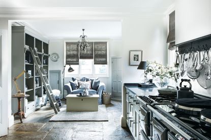 Kitchen with white walls and flagstone floor with black aga and gray cabinets with sitting area at far end and shelves with ladder