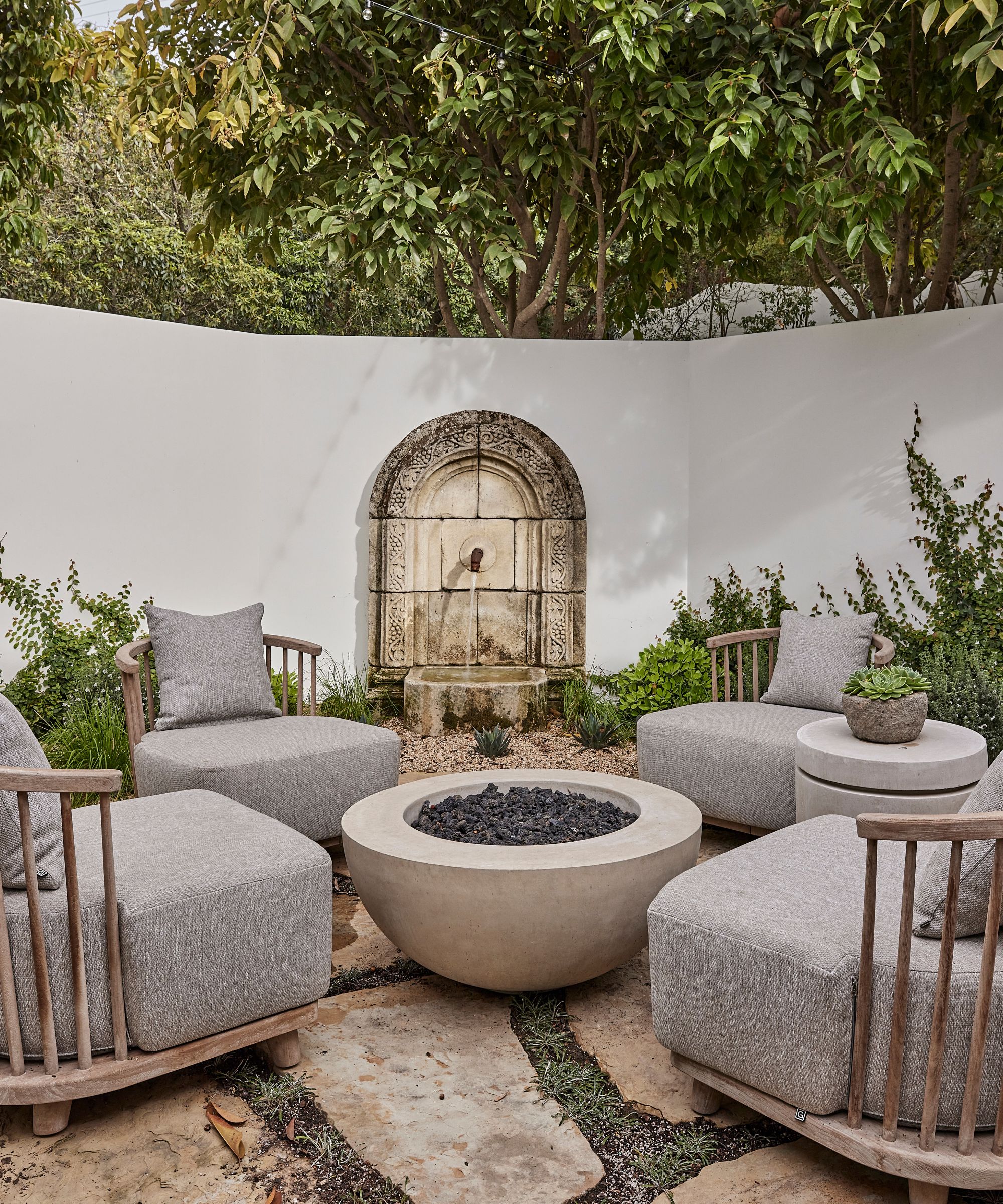 a natural spanish style outdoor sitting area with a stone fireplace, gray wooden chairs and an antique water feature with stone floors