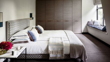 A neutral bedroom space with large brown closets, a window seat with cushions, and a large double bed with patterned headboard, white bedding, and cushions, and a marble bedside table beside it.