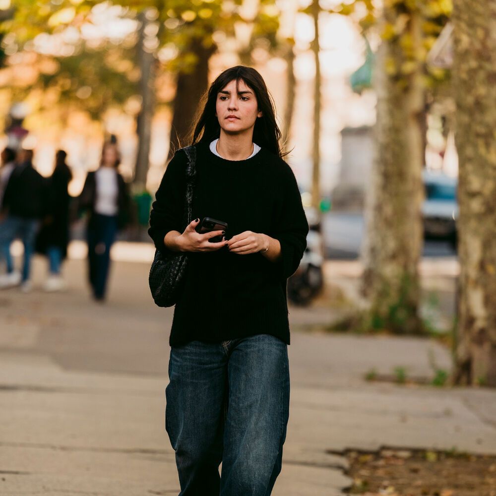 Woman wears relaxed black brew neck sweater with baggy dark wash jeans walking through the park. 