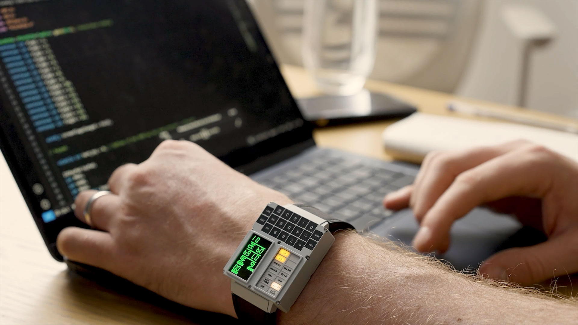 A pair of hands are shown working on a laptop computer. On the wrist of the left hand is a watch in the shape of the display and keypad (DSKY) for the Apollo Guidance Computer.