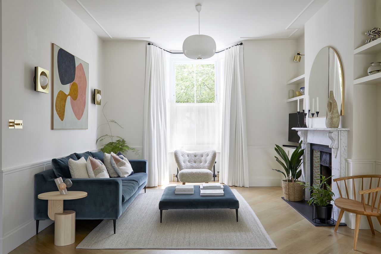 A living room with a wooden floor, fireplace, blue velvet sofa and long white sheer curtains in a bay window