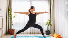 A woman performs a yoga flow at home. She wears a black vest top and black leggings and is on a blue yoga mat in front of a window.