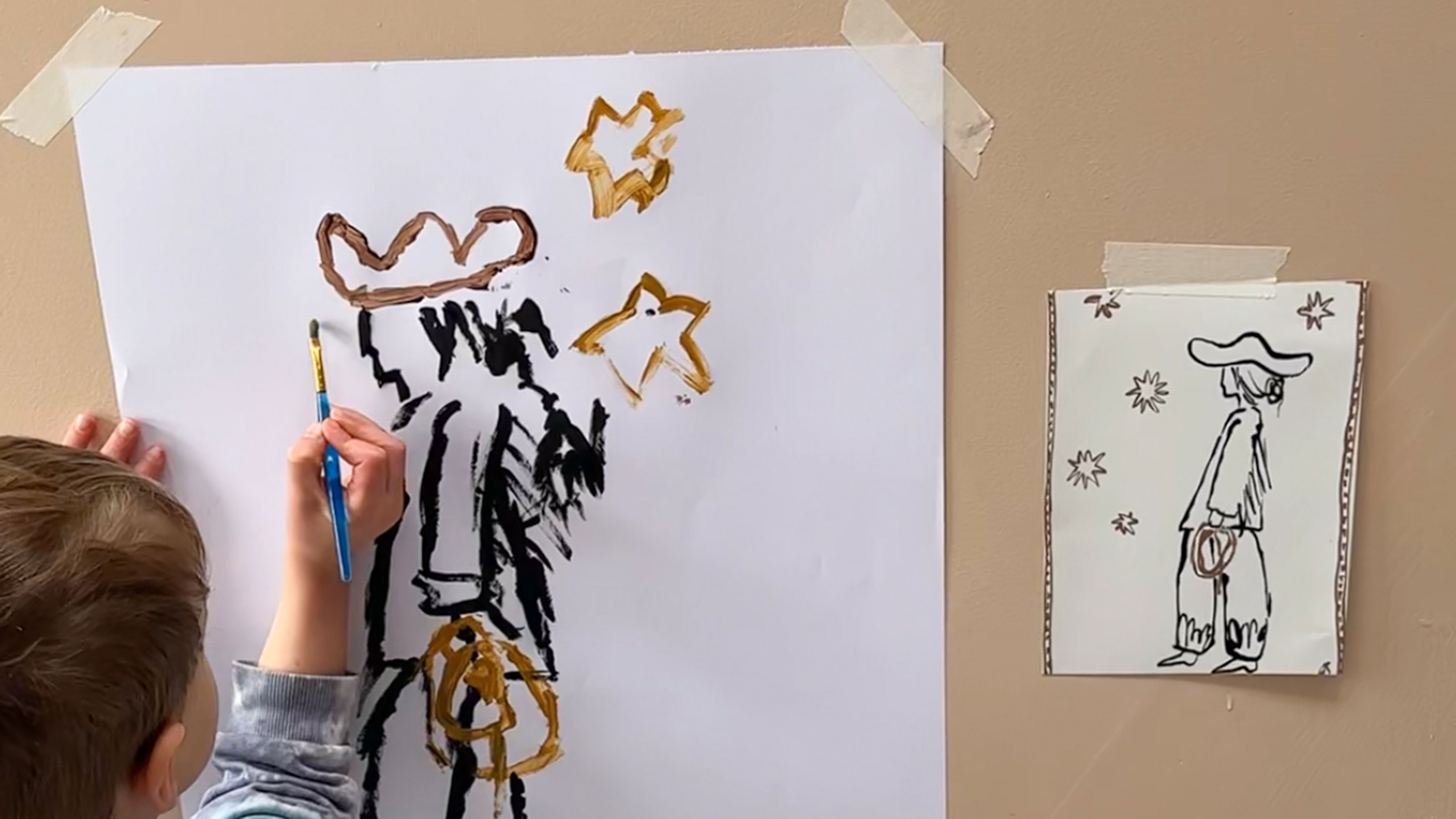Young child painting picture of cowboy surrounded by stars, coping picture on wall beside them
