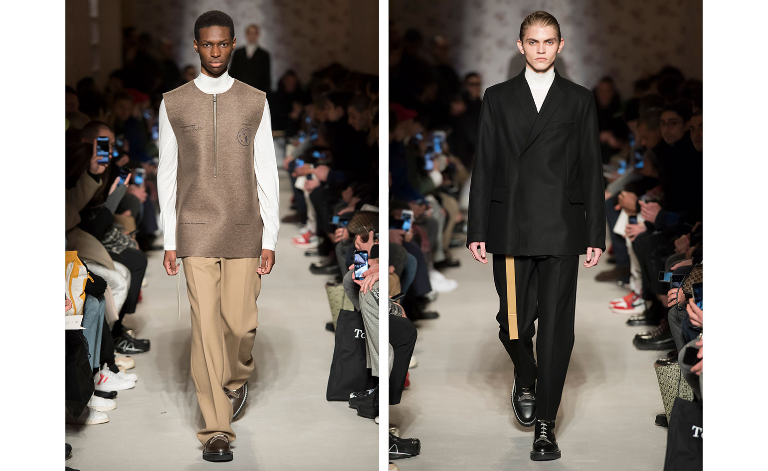 Catwalk view of two models wearing looks from OAMC's collection. One model is wearing a white high neck, long sleeve top, brown sleeveless piece, beige trousers and brown boots. And the second model is wearing a white high neck top, black suit, beige belt and black boots. There are people seated on both sides of the catwalk