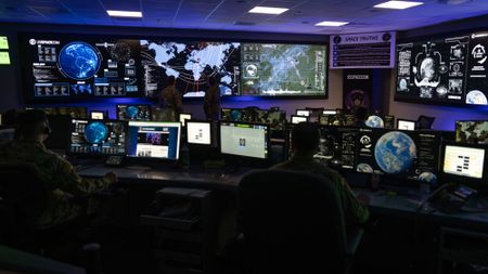 people in military fatigues sit behind computer consoles in a room full of screens showing earth and charts of satellites in orbit