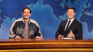 From left to right: Pete Davidson and Colin Jost sitting next to each other and smiling on SNL.