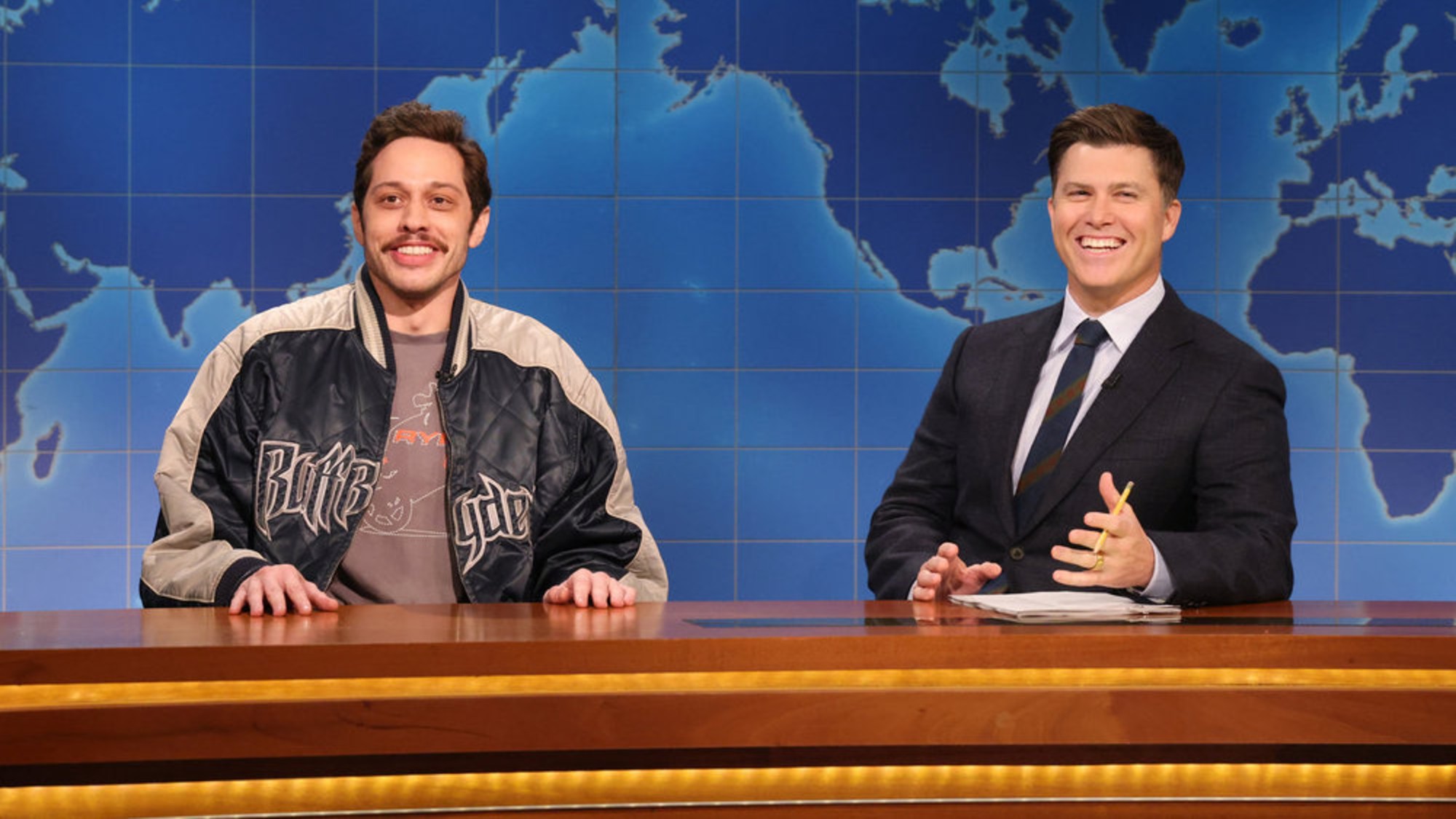 From left to right: Pete Davidson and Colin Jost sitting next to each other and smiling on SNL.
