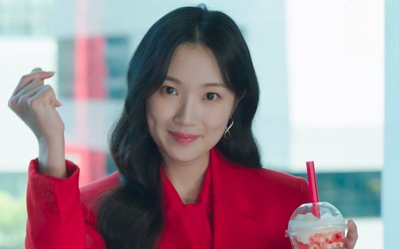 A young Korean woman (actress Kim Hye-yoon) wearing red makes a finger-snapping motion with her left hand as she holds a drink with whipped cream and red puree in her right. A still from the K-drama 'No Tail to Tell.'