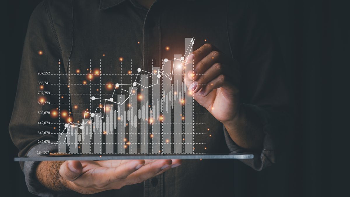 A photo of the hands and torso of a man wearing a dark shirt on a black background. His right hand is held palm up towards the camera at waist height with a tablet computer balanced on his fingertips. A holographic bar and line graph is springing from the tablet, and with his left hand he is highlighting part of the graph with a stylus.