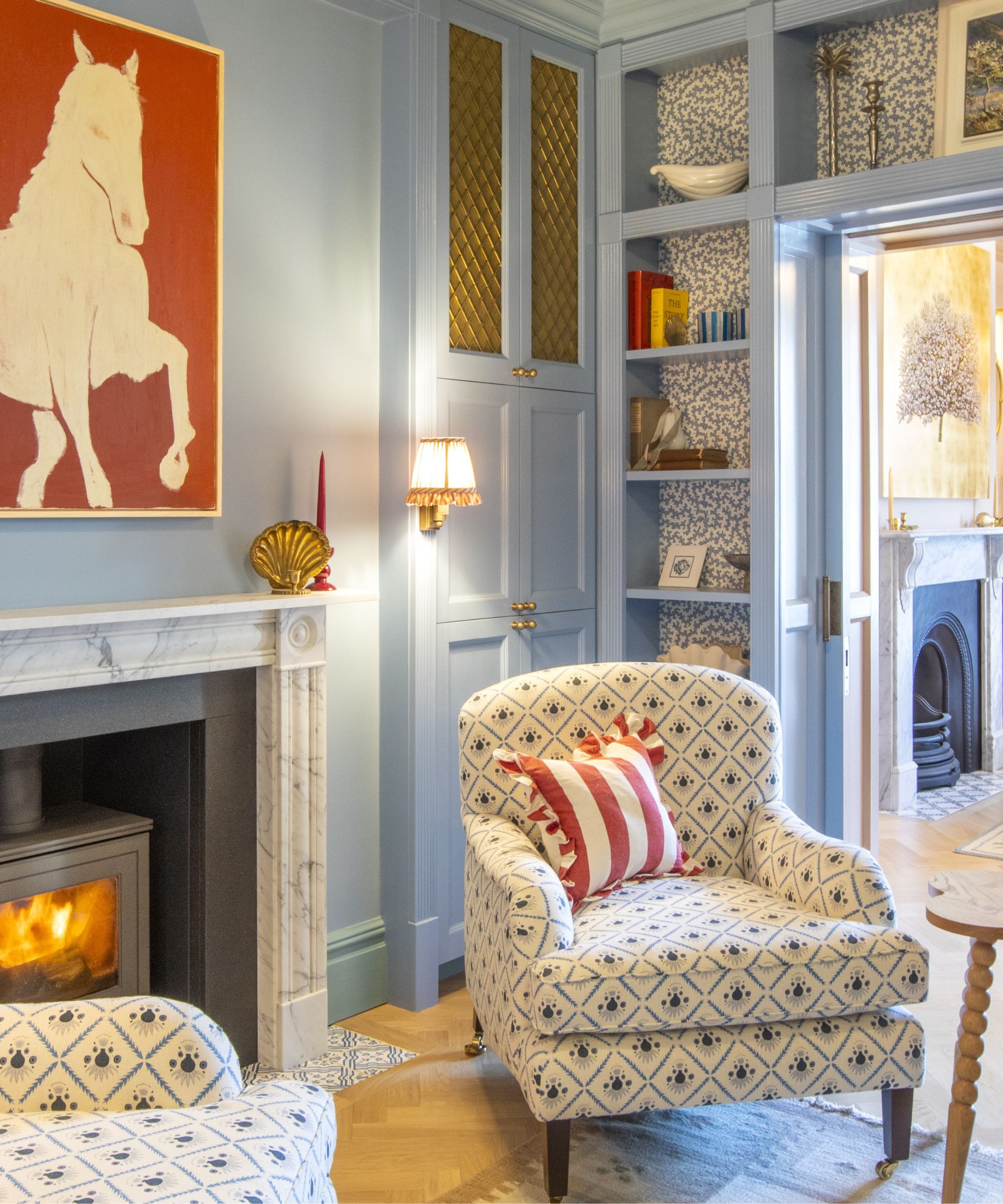 Blue living room with built-in wallpapered shelving, a marble fireplace with a log-burner, a large red painting of a horse above it, and a pair of cream floral armchairs on top of a vintage rug